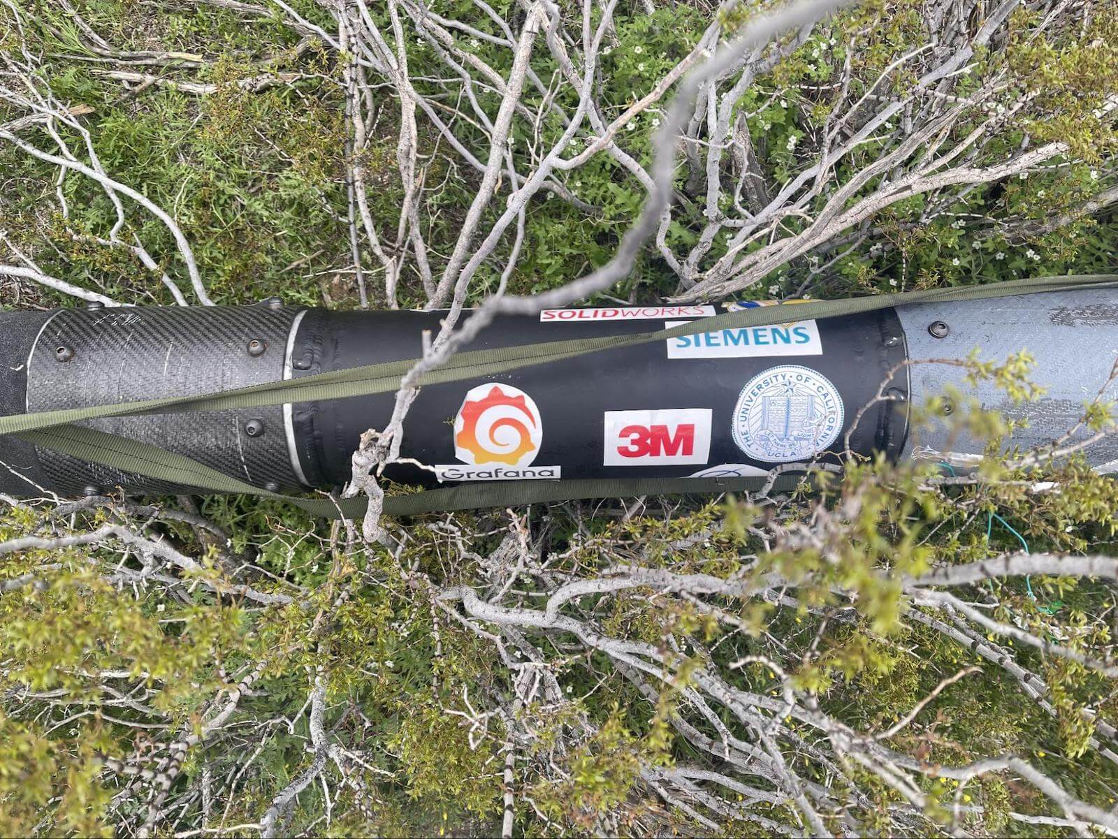 A photo of rocket in desert brush. The rocket is covered in stickers from Grafana, 3M, UCLA, Siemens, and Solidworks.