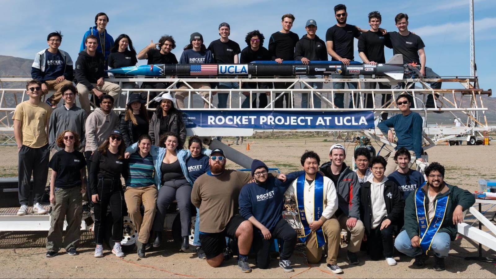 A photo of a group of people in the desert, holding a rocket and standing in front of a sign that says Rocket Project at UCLA