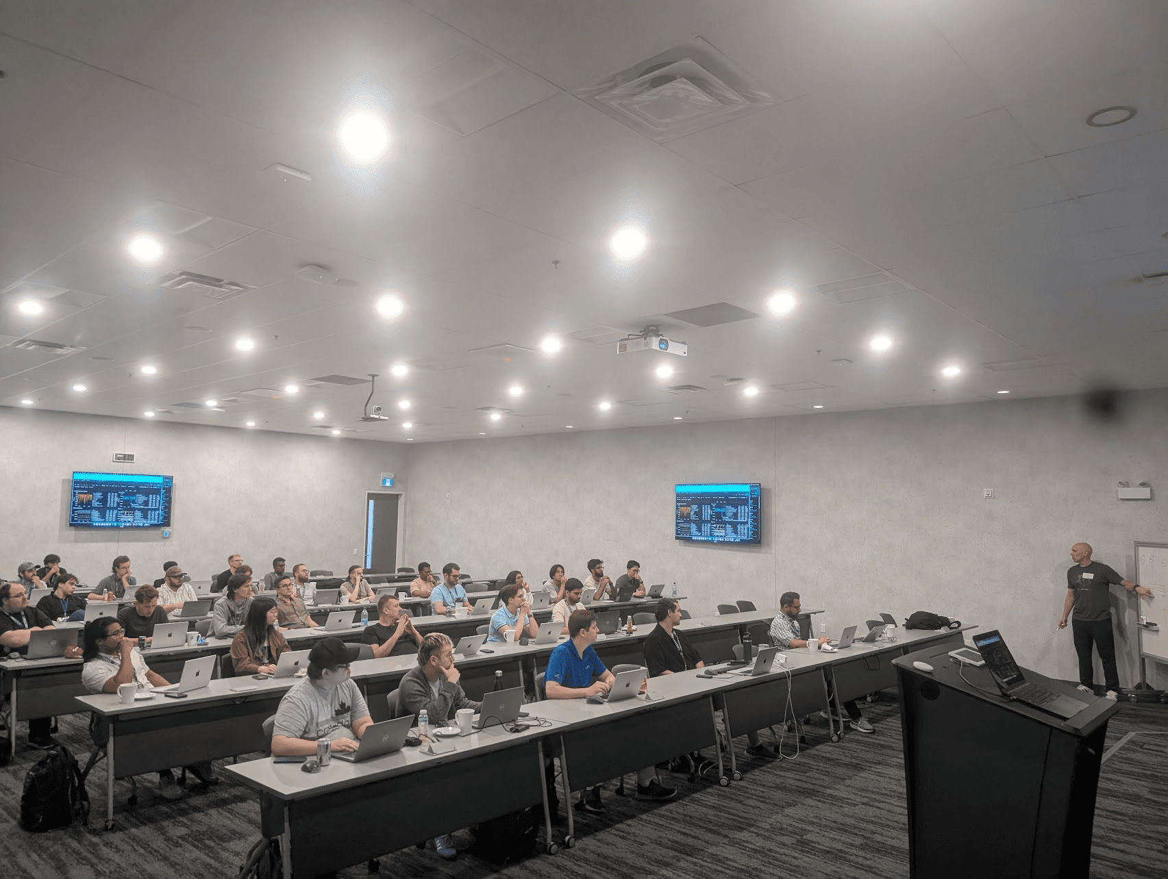 A room with rows of engineers seated at desks with laptops, listening to a presenter near a screen displaying a presentation.